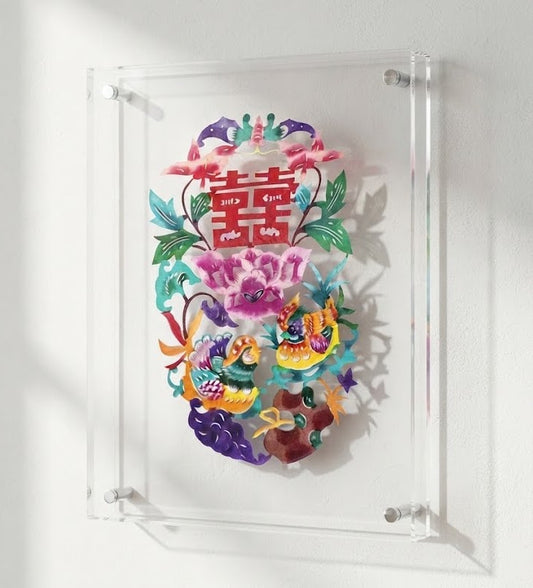 A colorful layered paper-cut wall art piece featuring a Chinese double happiness symbol, flowers, birds, and clouds, mounted in a clear acrylic frame against a white wall. Kaiwu studio